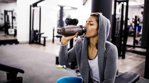 woman with a grey hoodie drinking a post workout shake in a gym